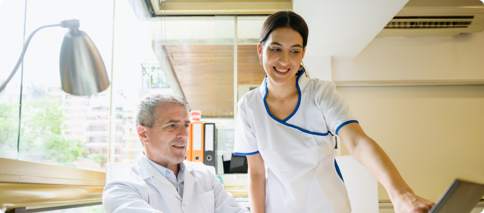 A doctor and nurse working together at a computer, smiling and discussing something on the screen. Image for the article 'How and when do we pay to a Professional.'